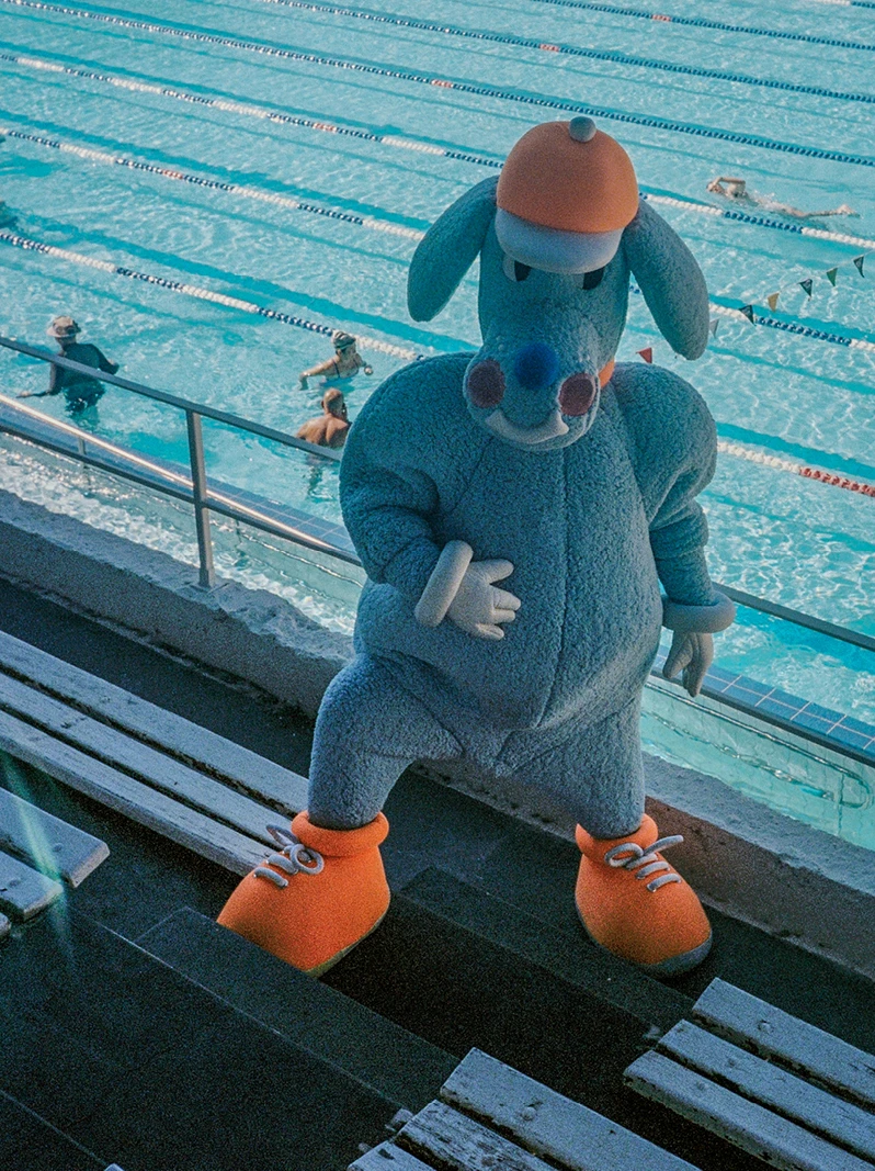 The blue Sports Dreams mascot standing on bleachers beside an outdoor swimming pool