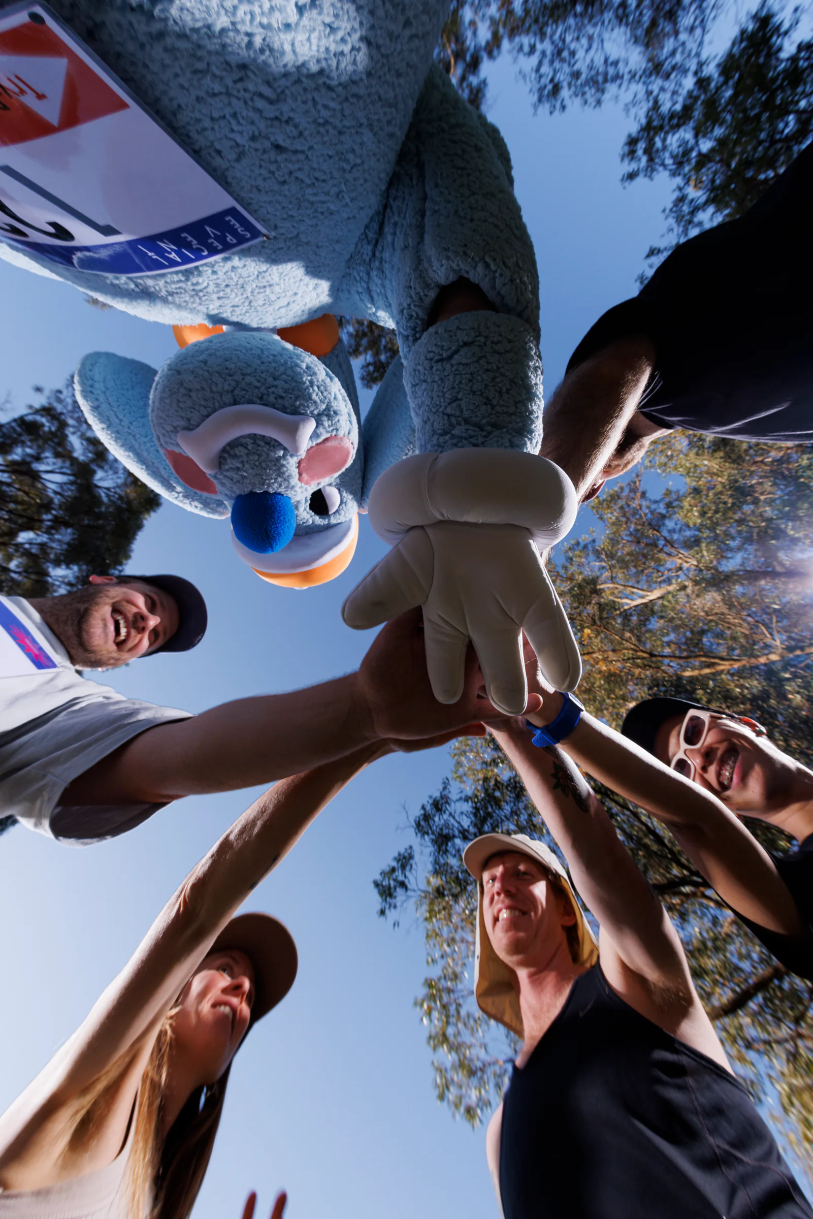 Low-angle team huddle with the blue Sports Dreams mascot, hands meeting beneath the sky